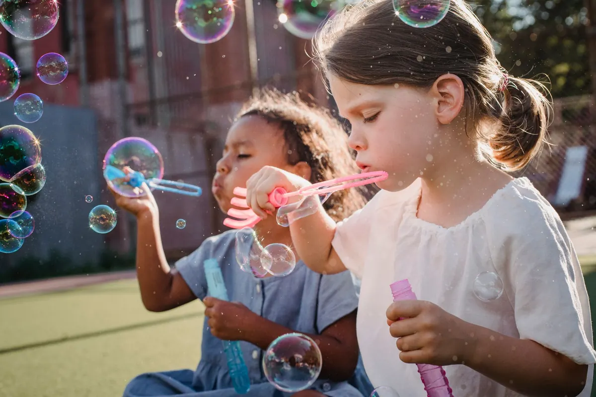 Two young girls joyfully blowing bubbles in the sunshine outdoors.