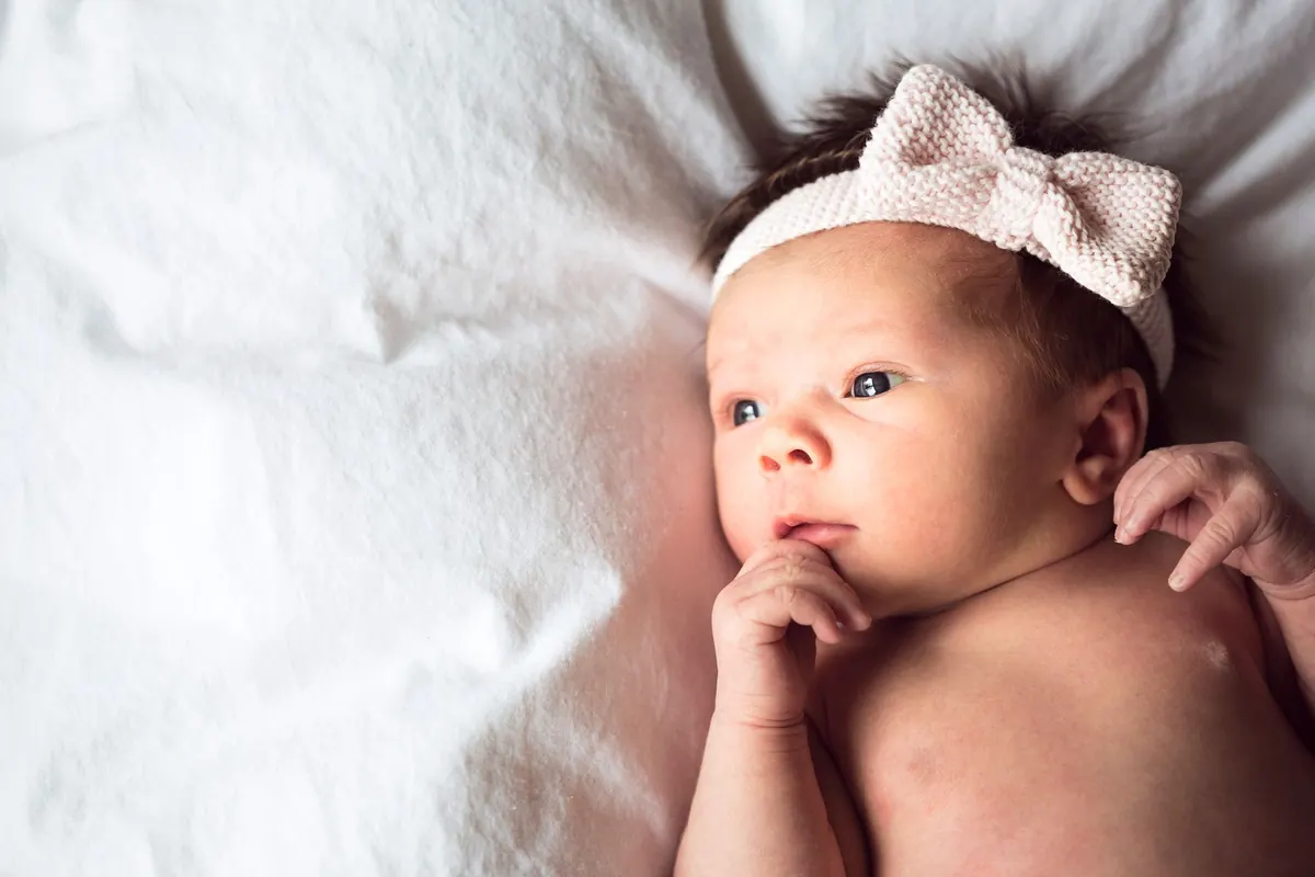 Charming portrait of a newborn baby with a pink bow headband lying on a soft white blanket.